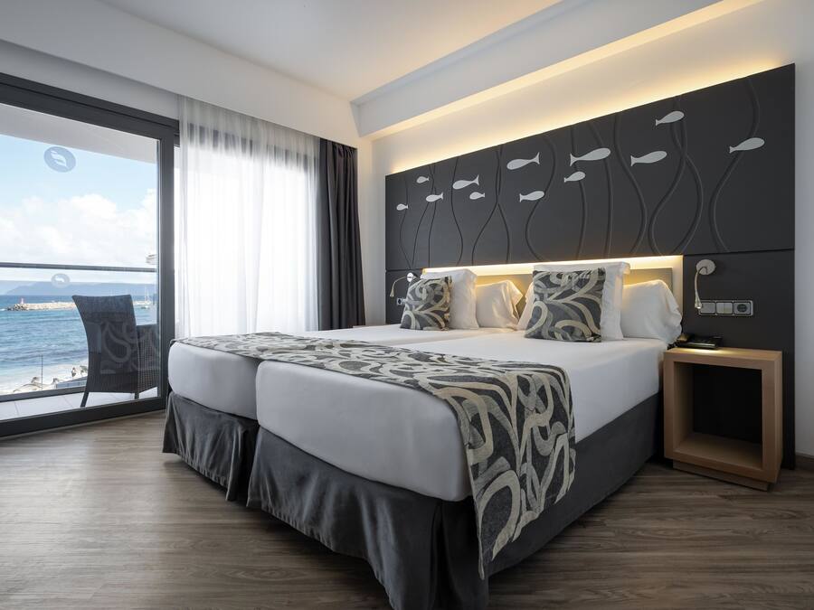 A modern hotel room with a large bed, a nightstand, and a view of the sea through a large window.