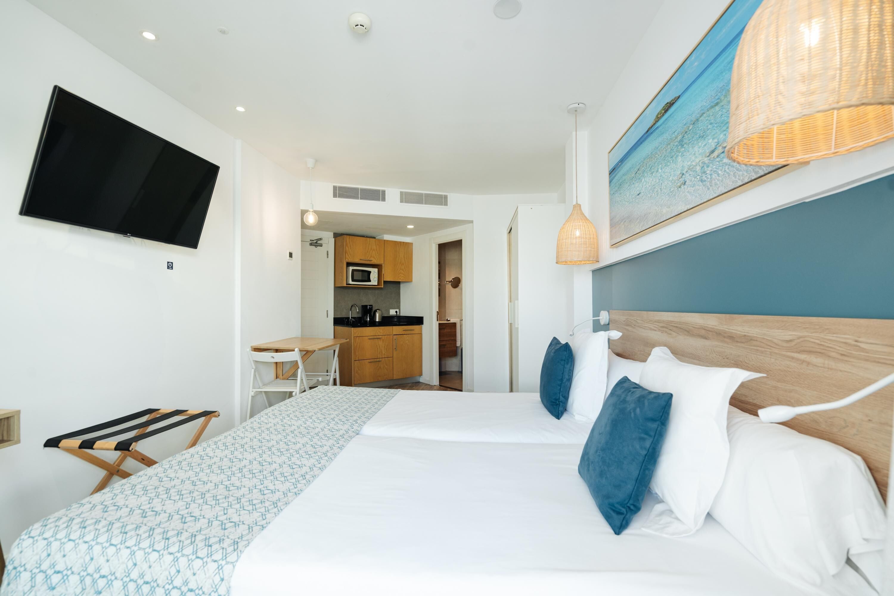 A modern hotel room with a large bed, a flat-screen TV mounted on the wall, a kitchen area, and a view of the sea.