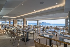 A spacious dining area with tables and chairs, large windows offering a view of the sea, and a modern ceiling design.