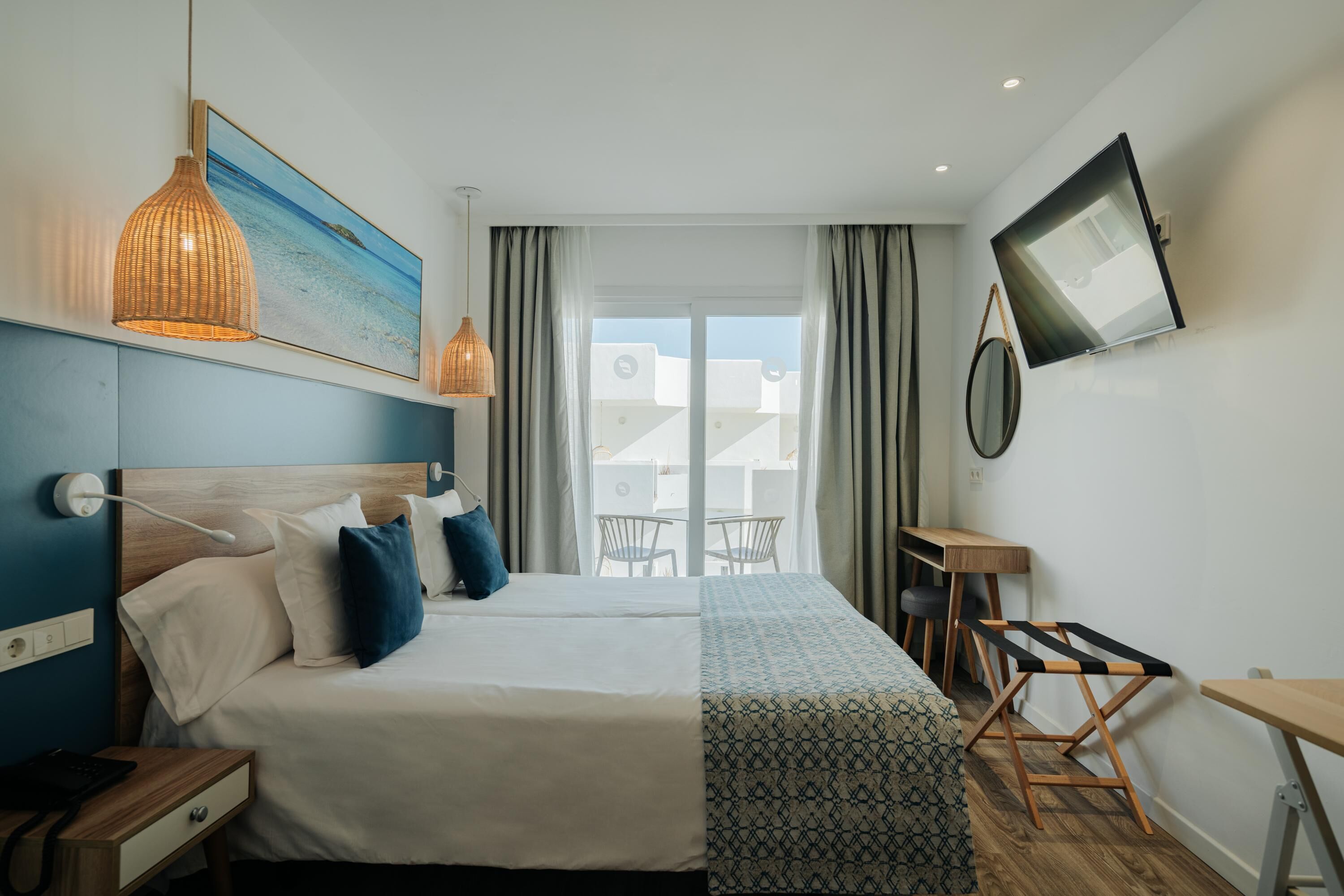 A hotel room with a bed, bedside table, lamp, TV, and a view of the outside.