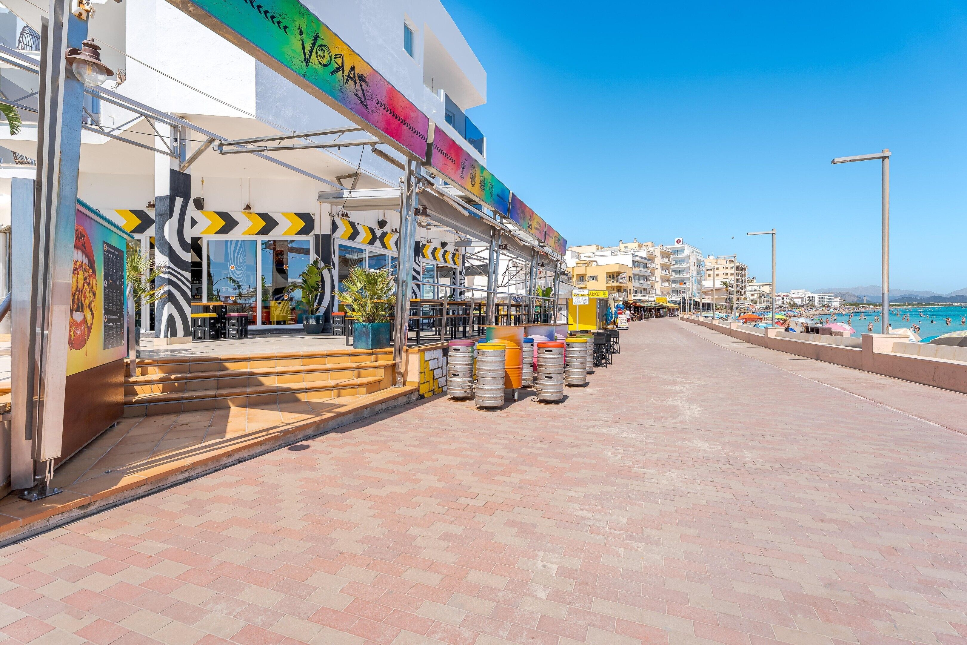 A seaside promenade with a restaurant featuring outdoor seating and a colorful awning.