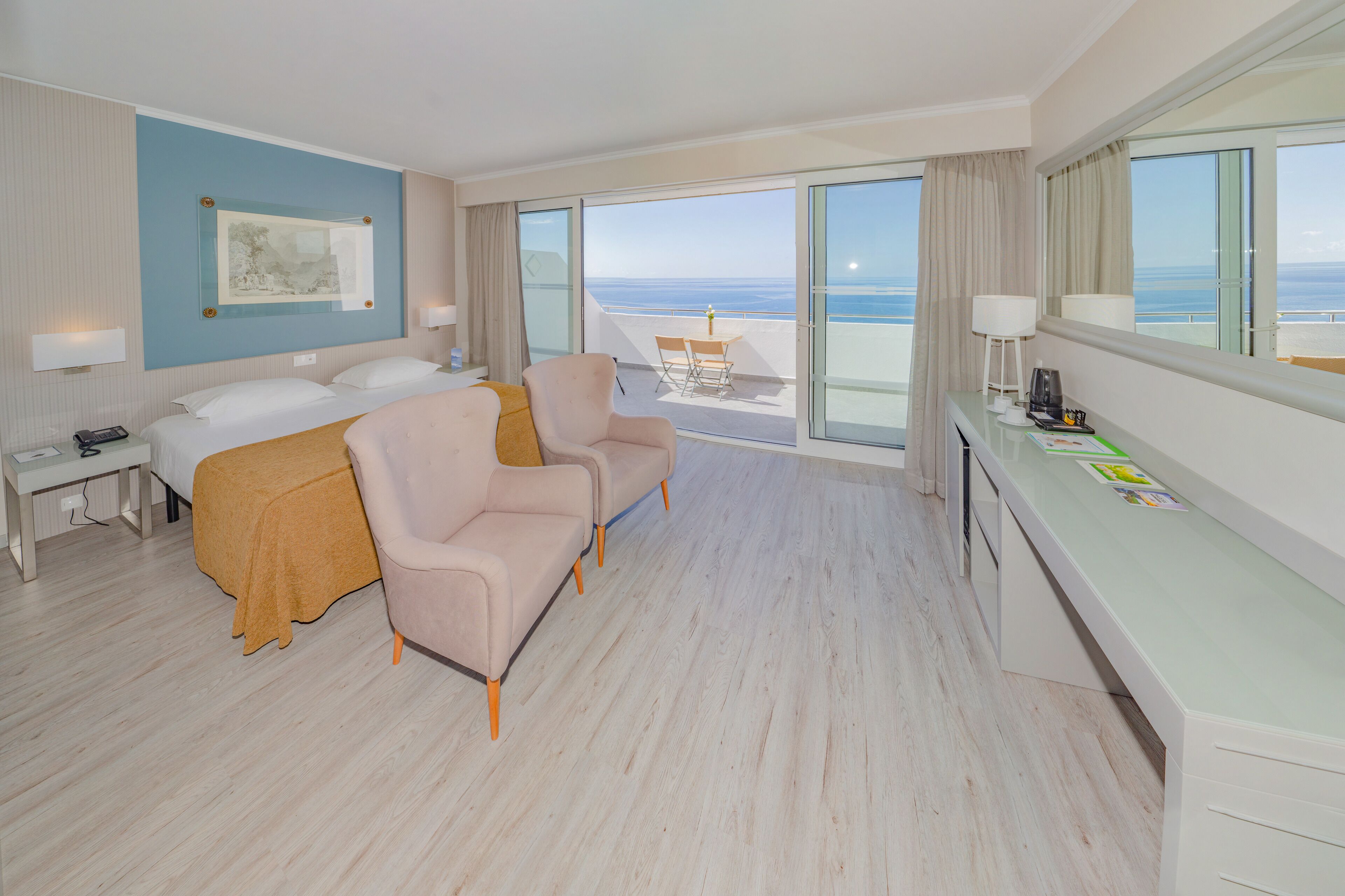 A hotel room with a bed, two chairs, a desk, and a view of the sea.