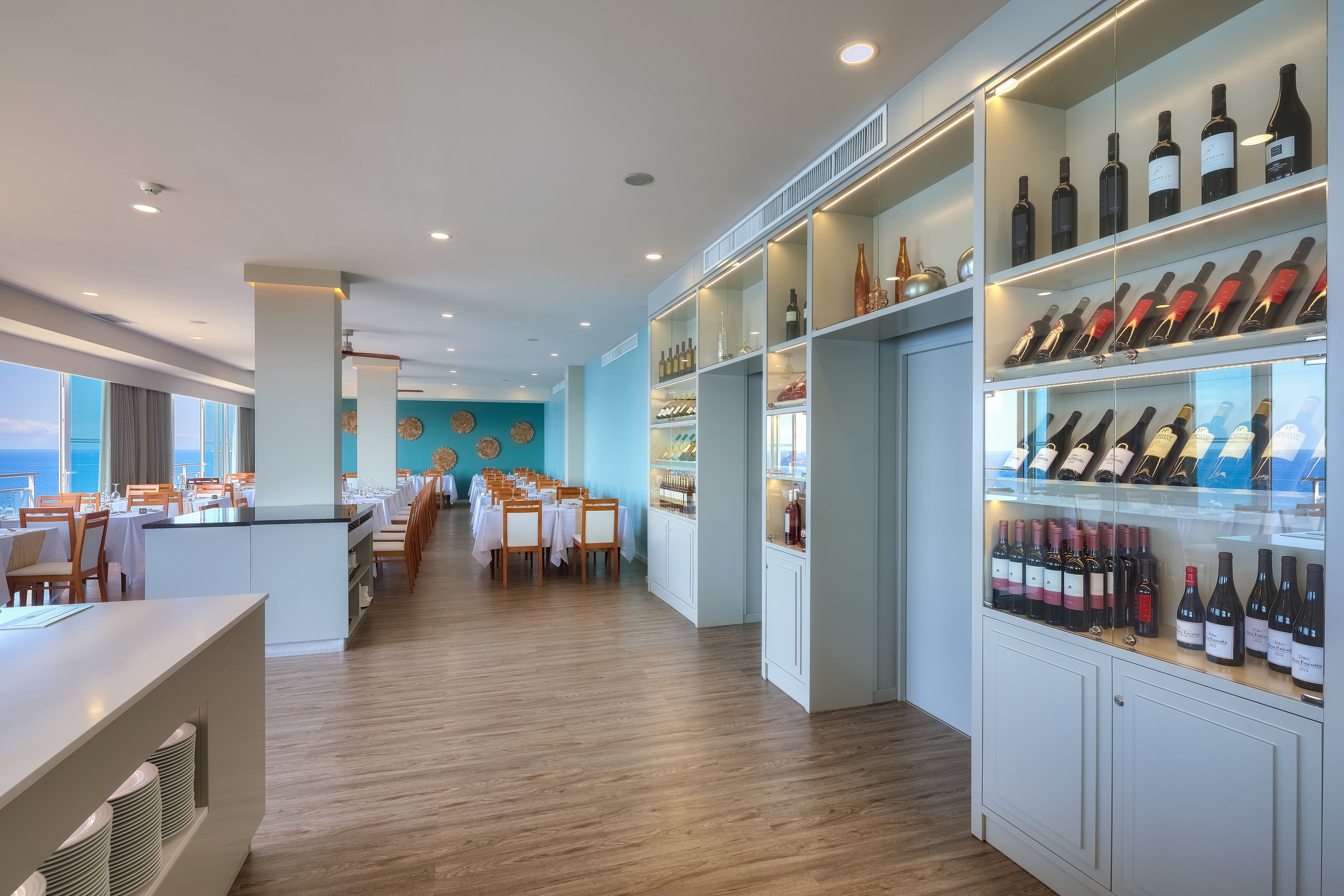 A restaurant with a long counter, multiple tables, and a wine display.