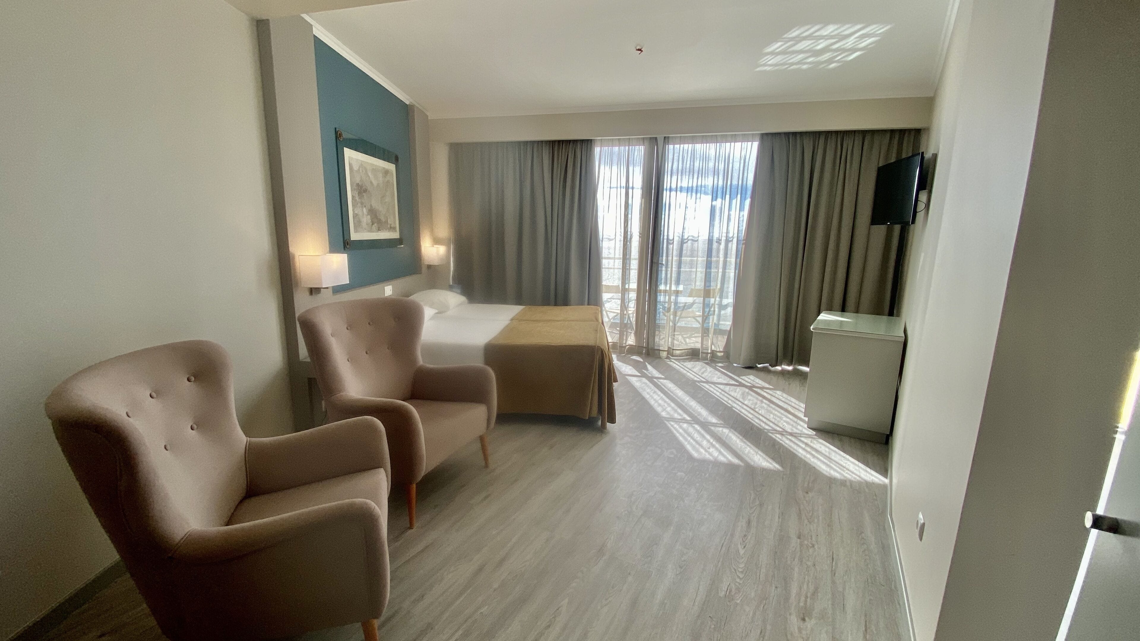 A hotel room with a bed, two armchairs, a small table, a TV, and a window with curtains.