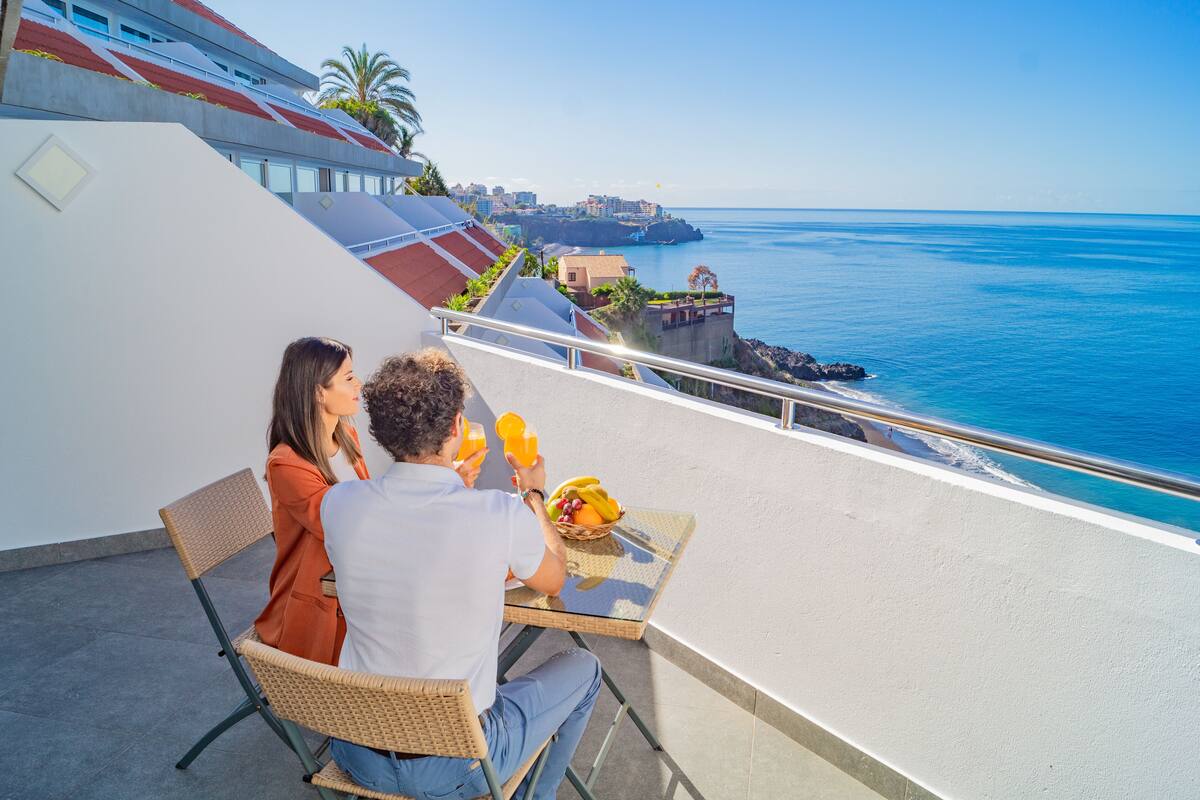 A couple dining on a balcony with a view of the sea and a coastal town.