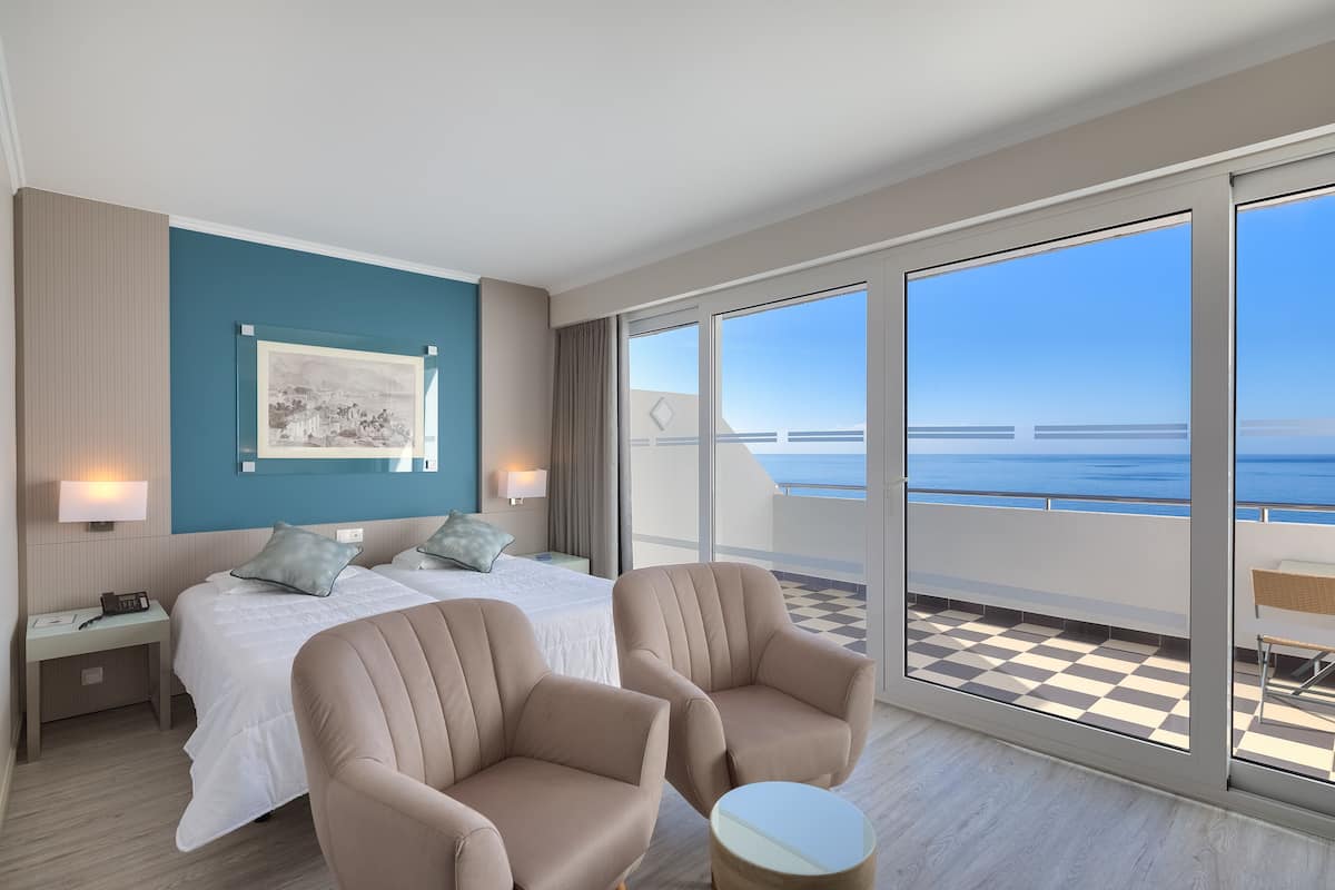 A hotel room with a large bed, two armchairs, a small table, and a balcony with a checkered floor and a view of the sea.