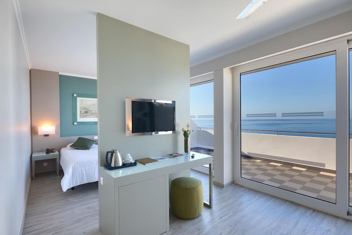 A modern hotel room with a bed, desk, and a large balcony with a view of the sea.