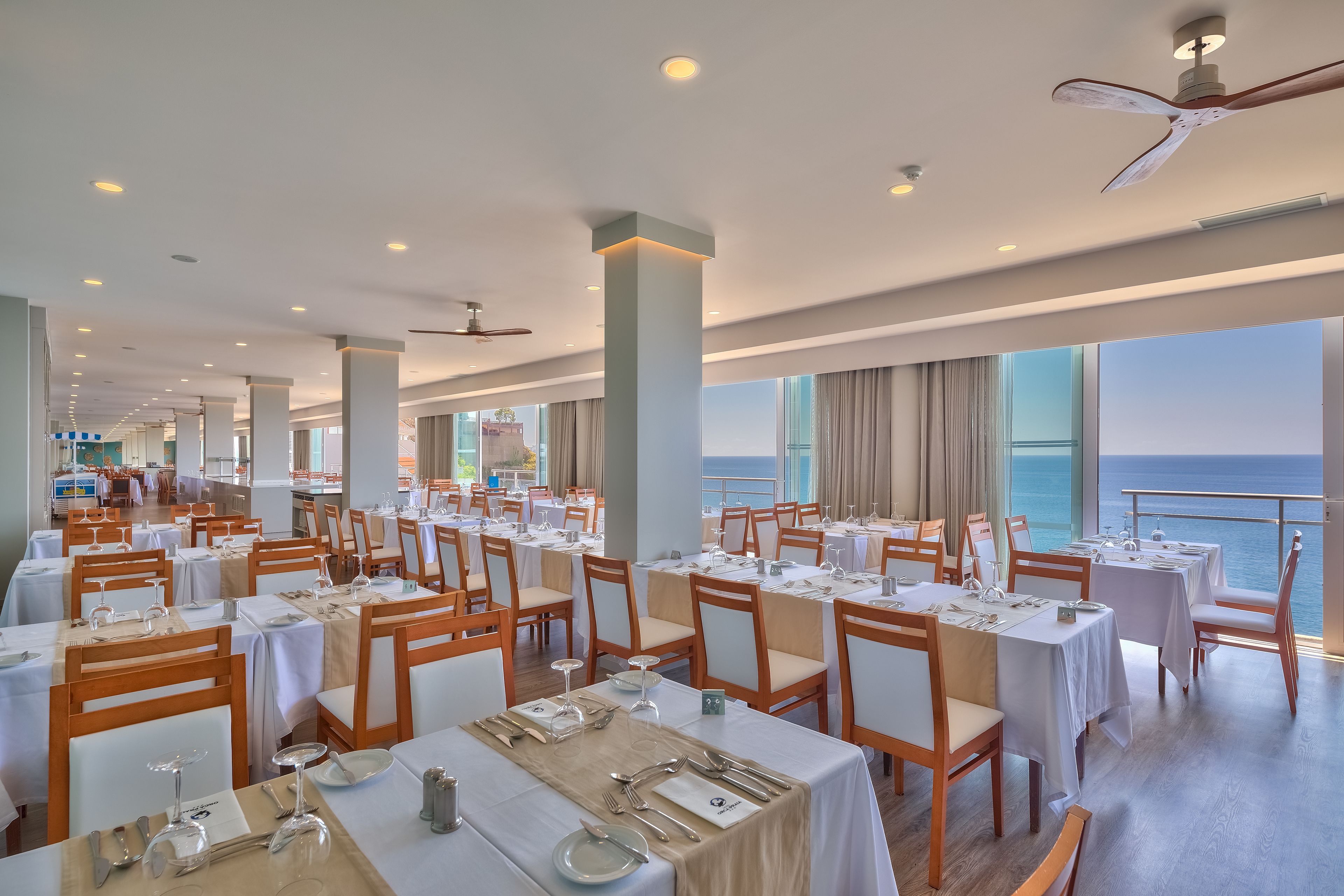 Un espace repas avec des tables dressées, de grandes fenêtres donnant sur la mer et un ventilateur de plafond.