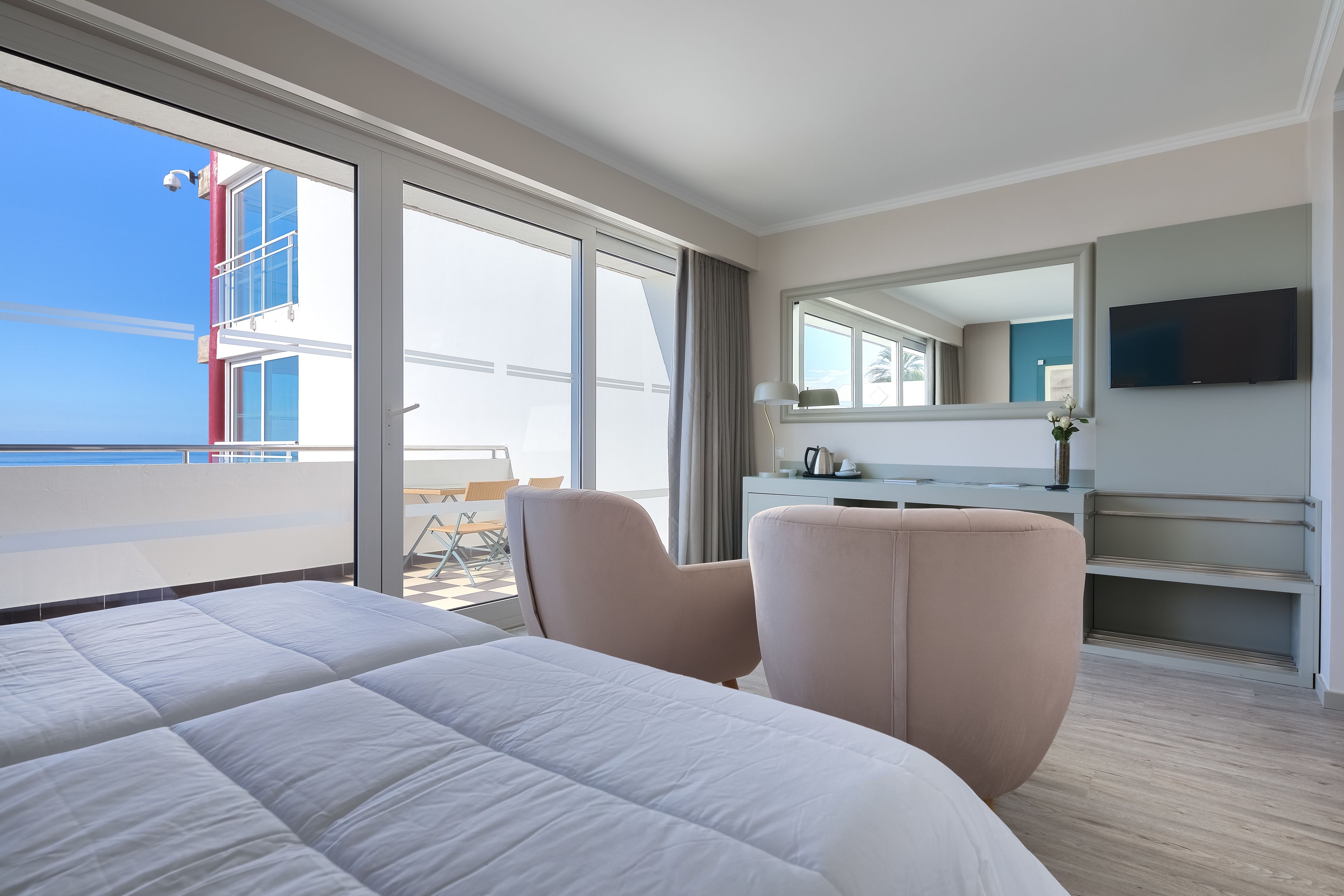 Habitación de hotel con una cama grande, zona de estar con una silla, escritorio, televisor de pantalla plana y balcón con vistas al edificio y al cielo.
