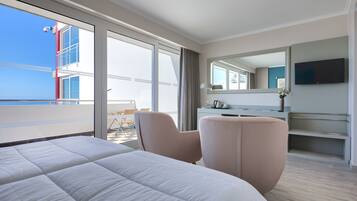 Habitación de hotel con una cama grande, zona de estar con una silla, escritorio, televisor de pantalla plana y balcón con vistas al edificio y al cielo.