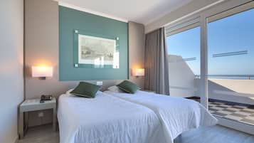 A hotel room with two beds, a nightstand, a lamp, a framed picture on the wall, and a view of the sea through a large window.