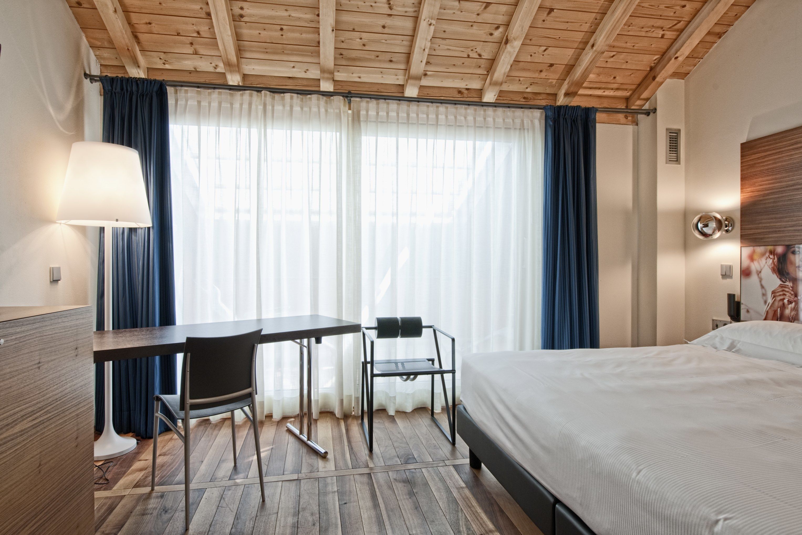 Ett hotellrum med ett träpanelat tak, en säng, ett skrivbord med en stol och ett fönster med gardiner.