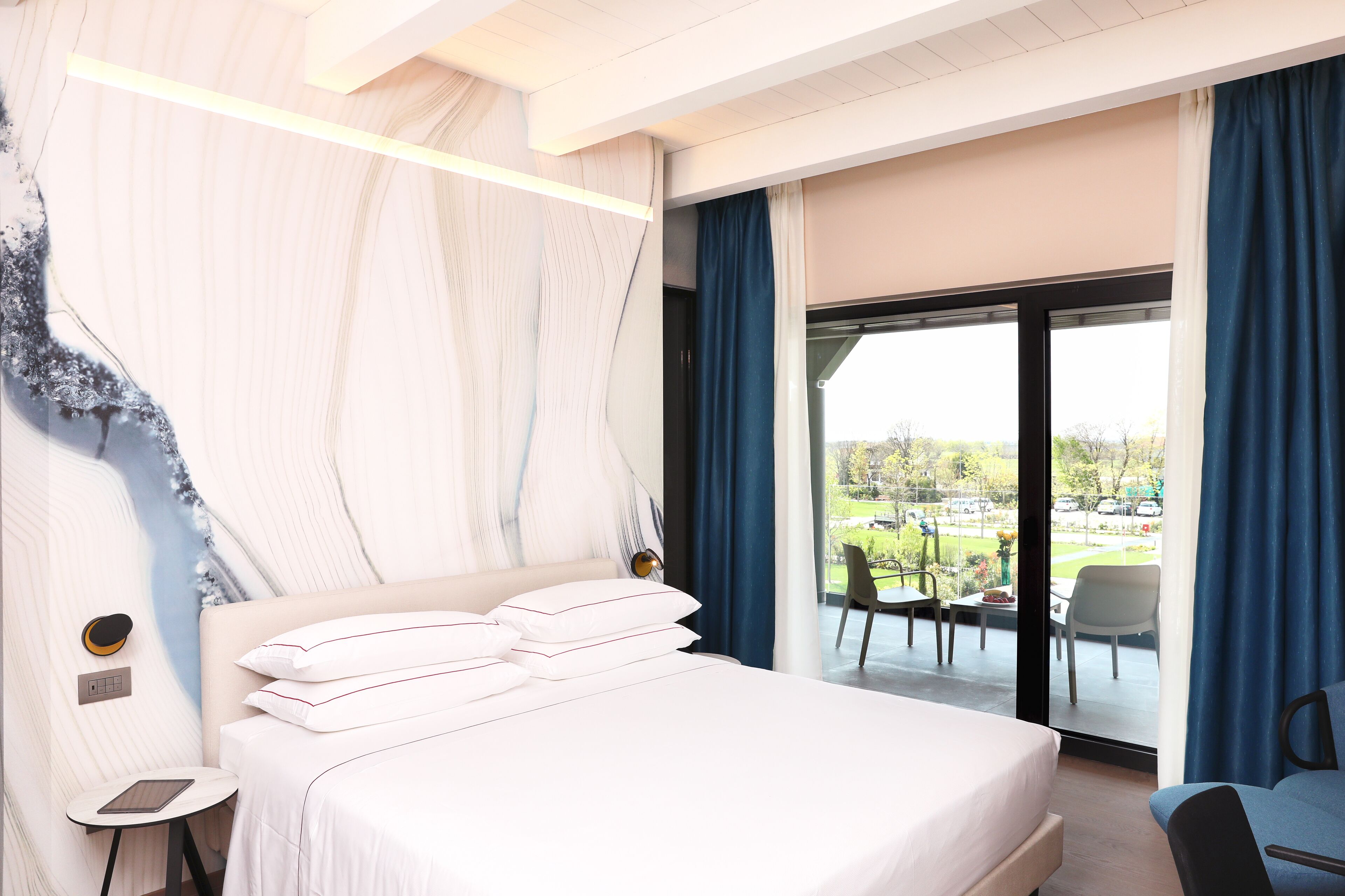 A modern hotel room with a large bed, a bedside table, a chair, and a view of the outdoors.