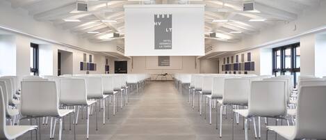 A conference room with rows of white chairs, a projector screen, and a ceiling with exposed beams and lighting fixtures.