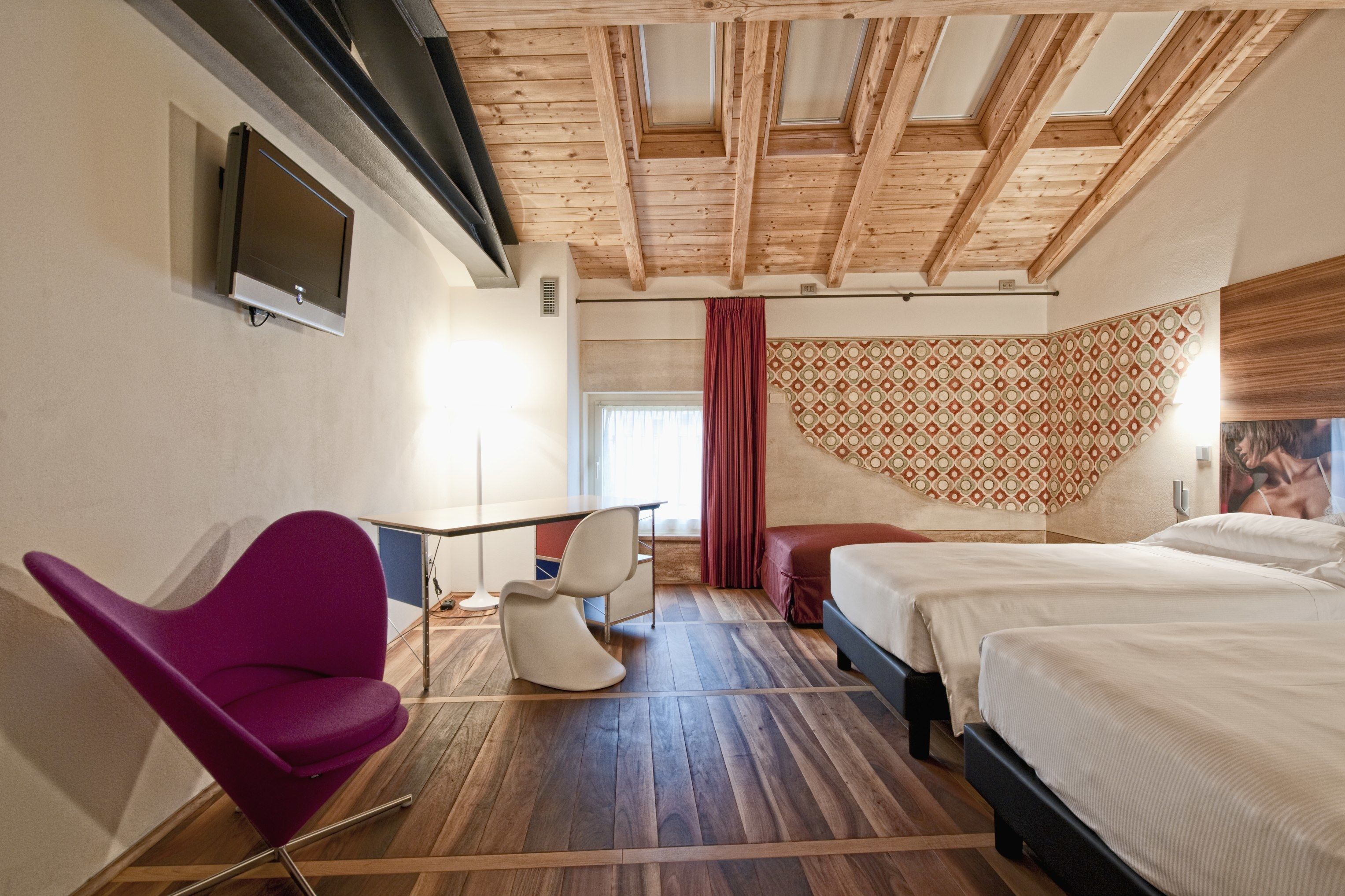 Habitación de hotel con techo de madera, televisor montado en la pared, escritorio con sillas, cama y pared con diseño.