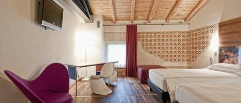 A hotel room with a wooden ceiling, a TV mounted on the wall, a desk with chairs, a bed, and a patterned wall.
