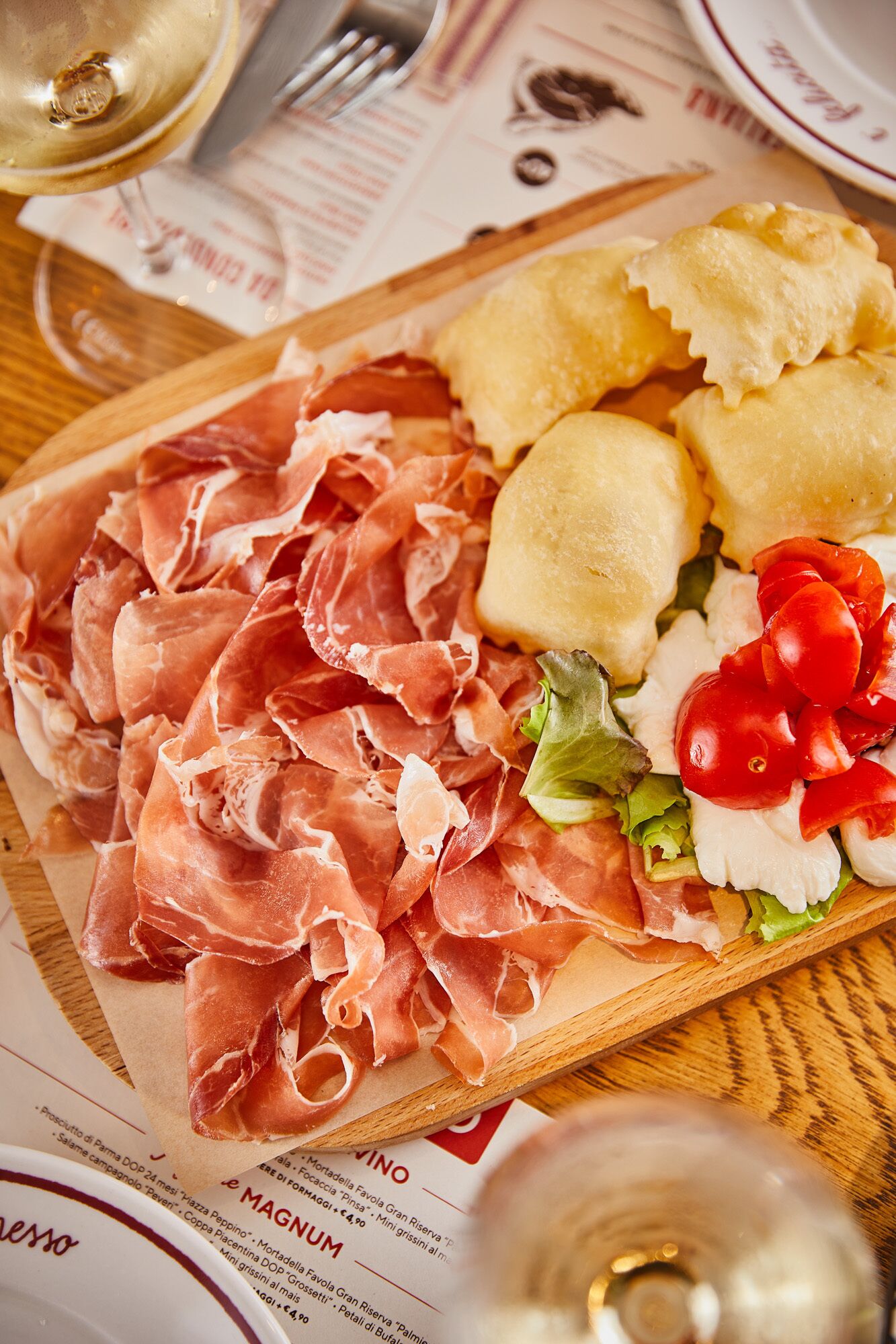 A wooden board with sliced prosciutto, bread, and a salad with tomatoes and greens.