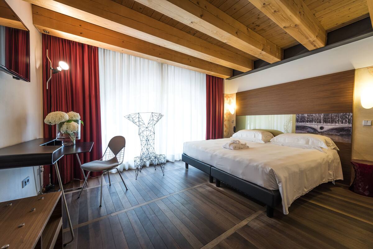 A hotel room with a wooden ceiling, a bed with white bedding, a desk with a chair, and a large window with red curtains.