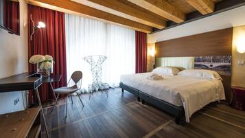 Quarto de hotel com teto de madeira, cama com roupa de cama branca, uma escrivaninha com cadeira e uma janela grande com cortinas vermelhas.