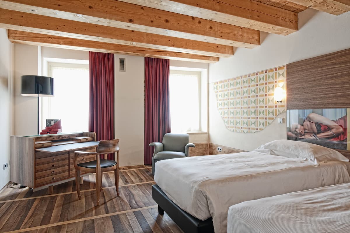 A hotel room with a wooden ceiling, a desk with drawers, a chair, a bed with white bedding, a window with red curtains, and a wall-mounted light fixture.