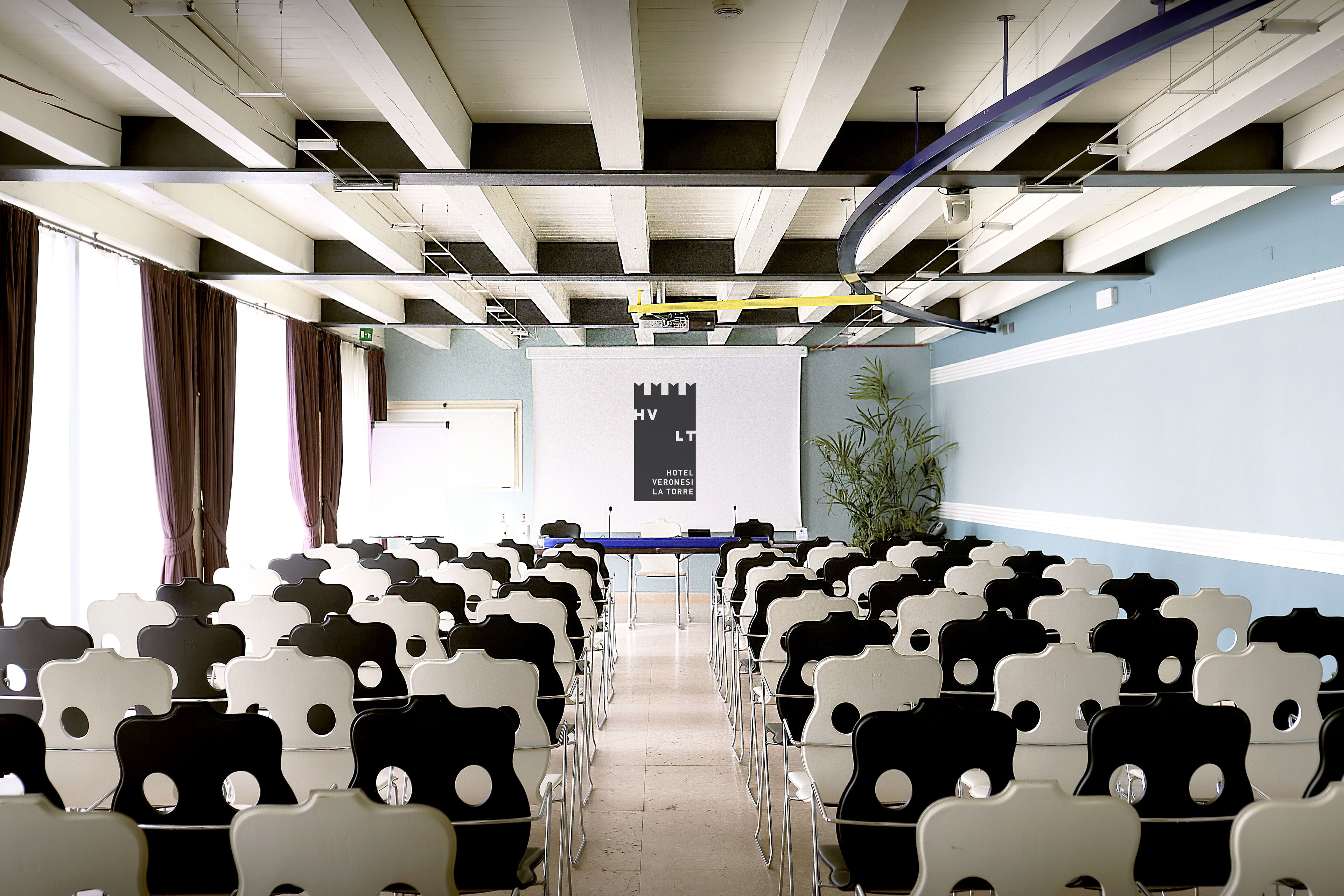 Uma sala de conferências com fileiras de cadeiras voltadas para uma tela de apresentação, uma planta à direita e uma escada ao fundo.