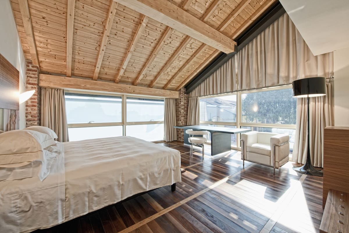 A bedroom with a large bed, a desk, and a chair, wooden flooring, and a view through a large window.