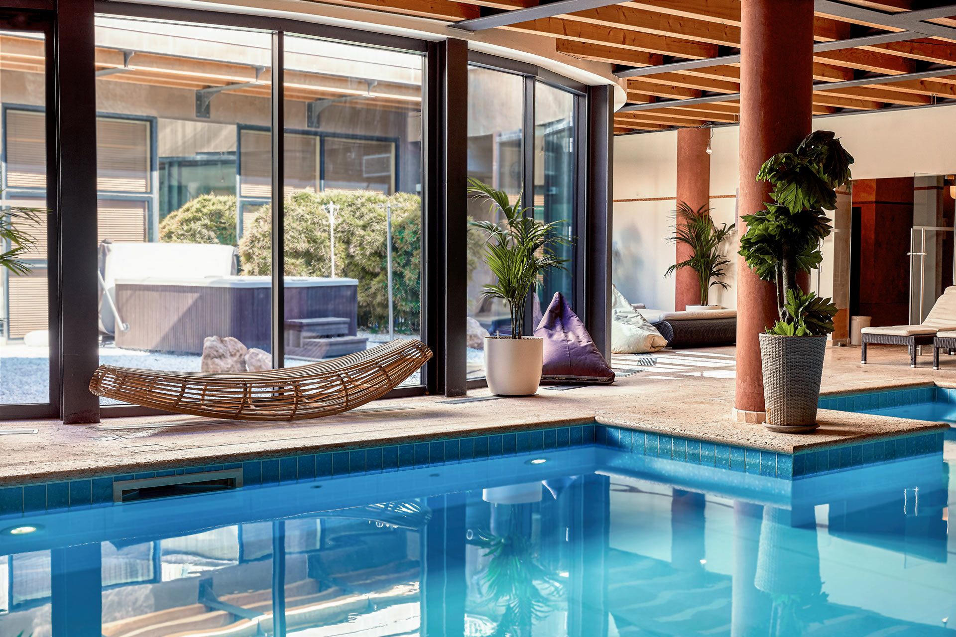 A modern indoor pool area with clear glass walls, a hot tub, and lounge chairs.