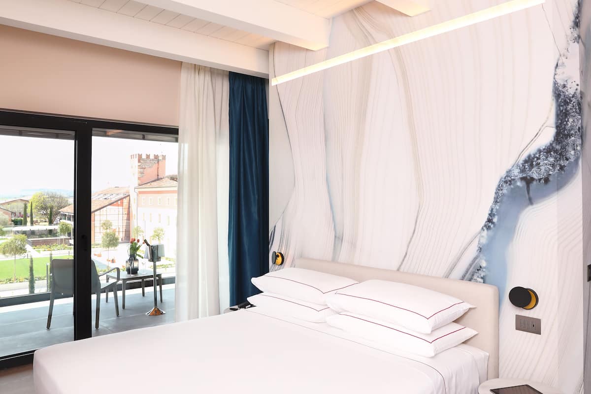 A modern hotel room with a large bed, a view of the city, and a balcony with a table and chairs.