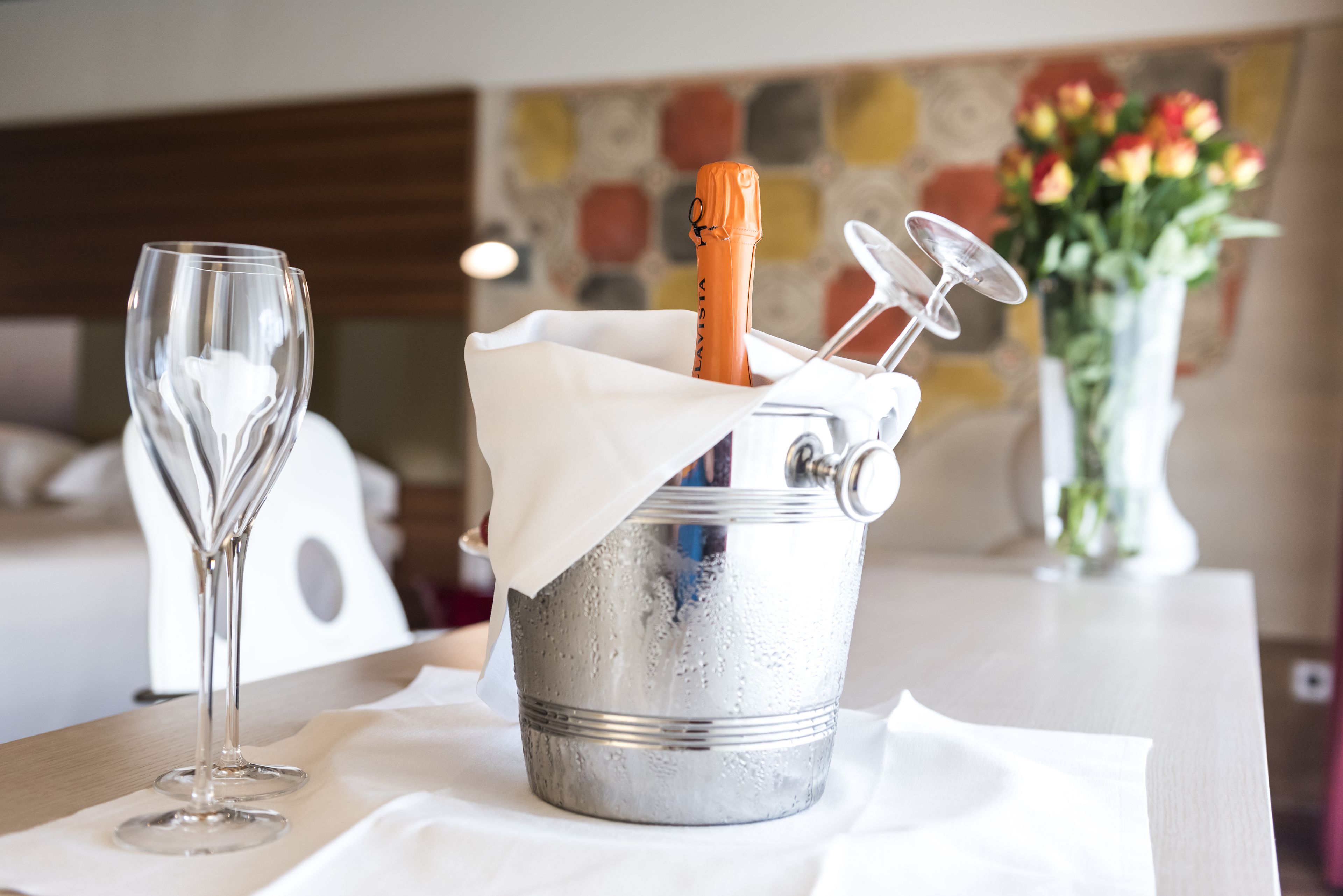 A champagne bottle in a metal bucket with a foil wrapper, a wine glass, and a vase of flowers on a table.