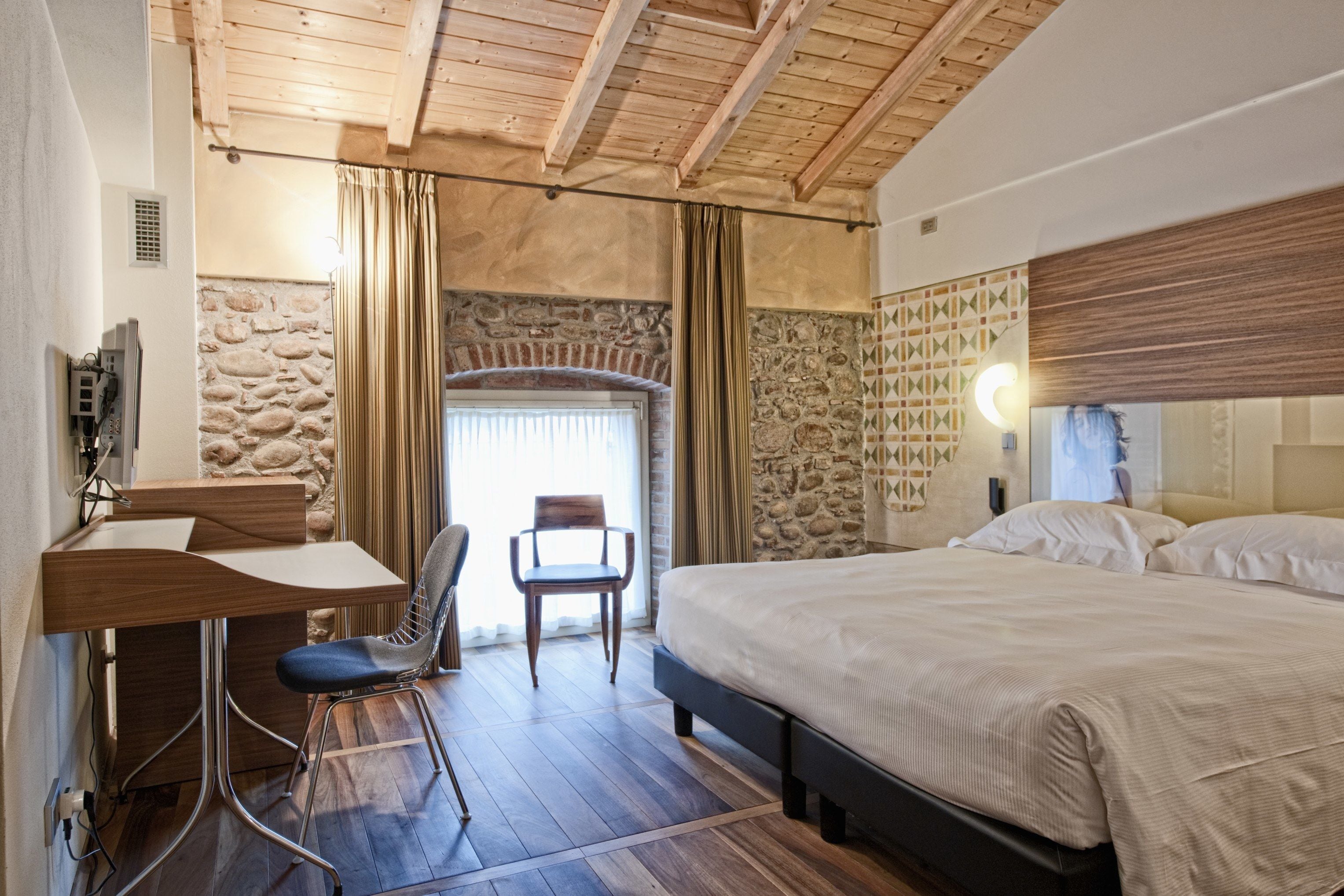 Ein Schlafzimmer mit Holzboden, Steinwand, einem Bett, einem Schreibtisch mit Stuhl und einem fenster mit Vorhängen.