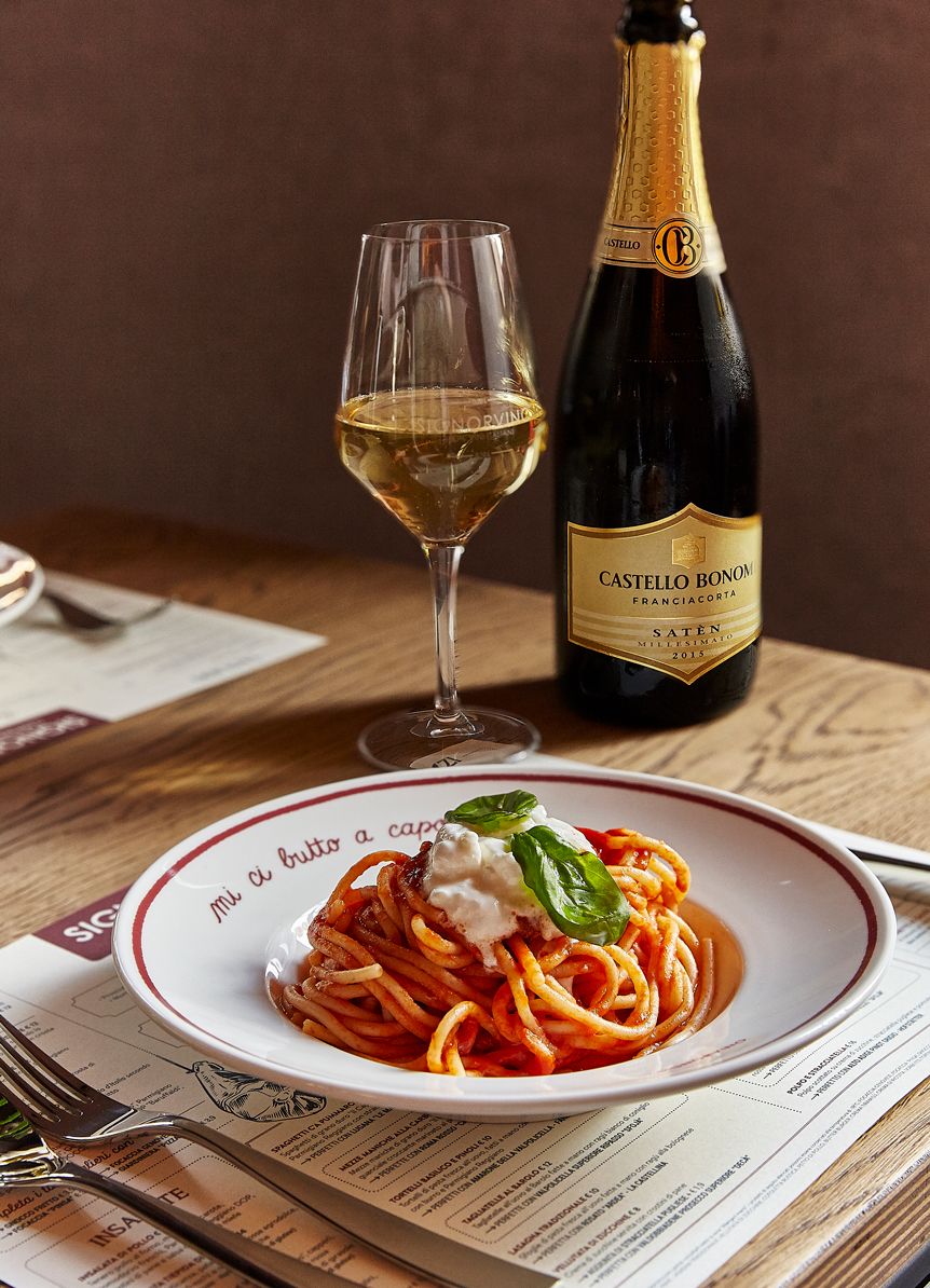 A plate of spaghetti with a creamy topping and basil, a glass of white wine, and a bottle of Castello Bonom wine on a wooden table.