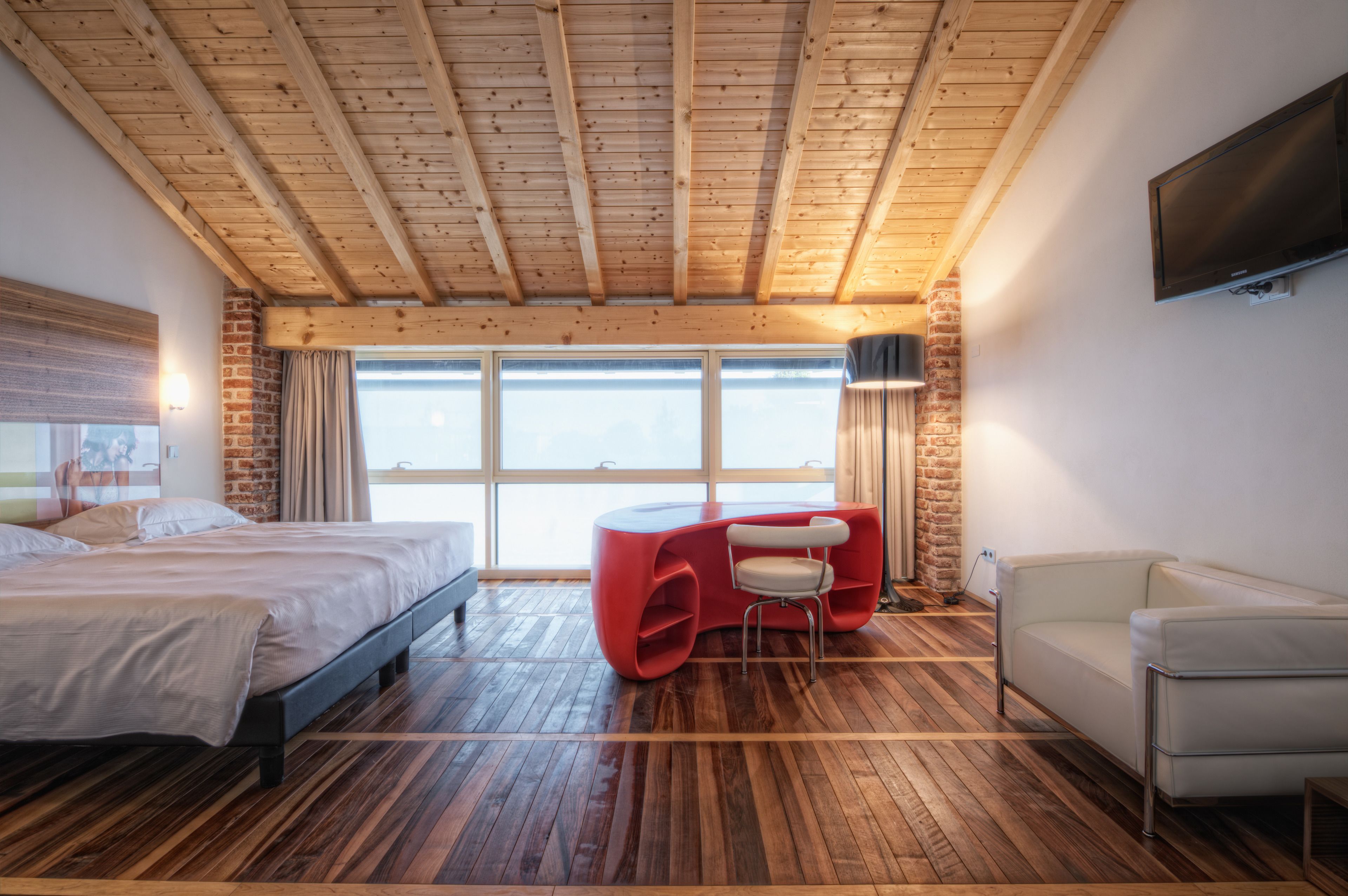 Een moderne slaapkamer met een houten plafond, een groot bed, een rode zithoek en een aan de muur bevestigde televisie.
