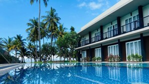 A swimming pool with clear blue water, surrounded by palm trees and a hotel building with balconies.