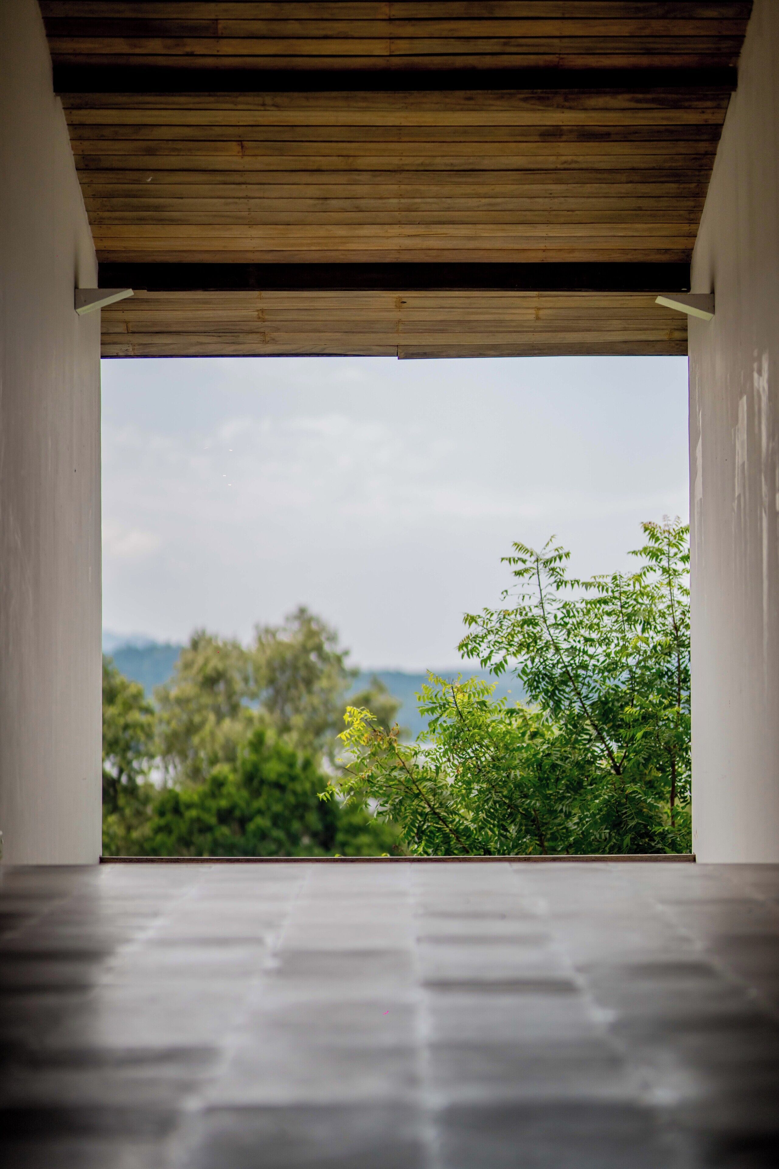 A room with a concrete floor and a wooden ceiling, leading to a view of greenery and a distant landscape.