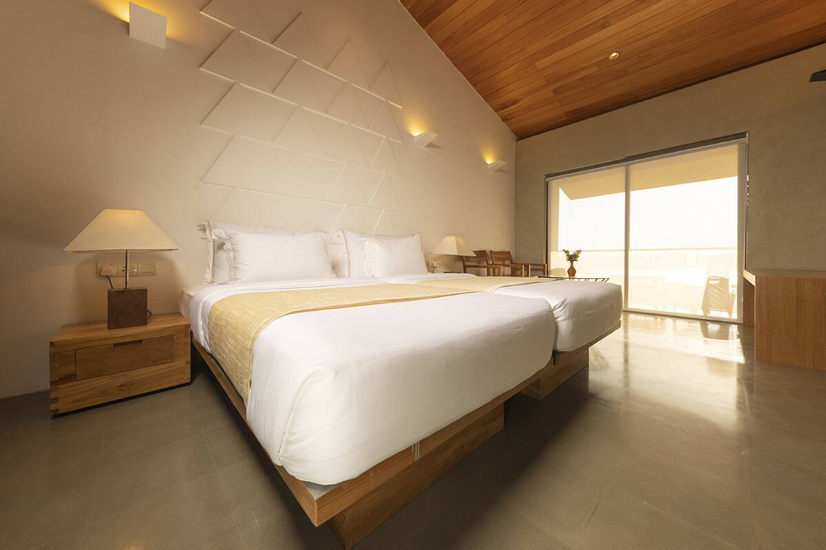A modern bedroom with two beds, wooden headboard, bedside tables, and a large window.