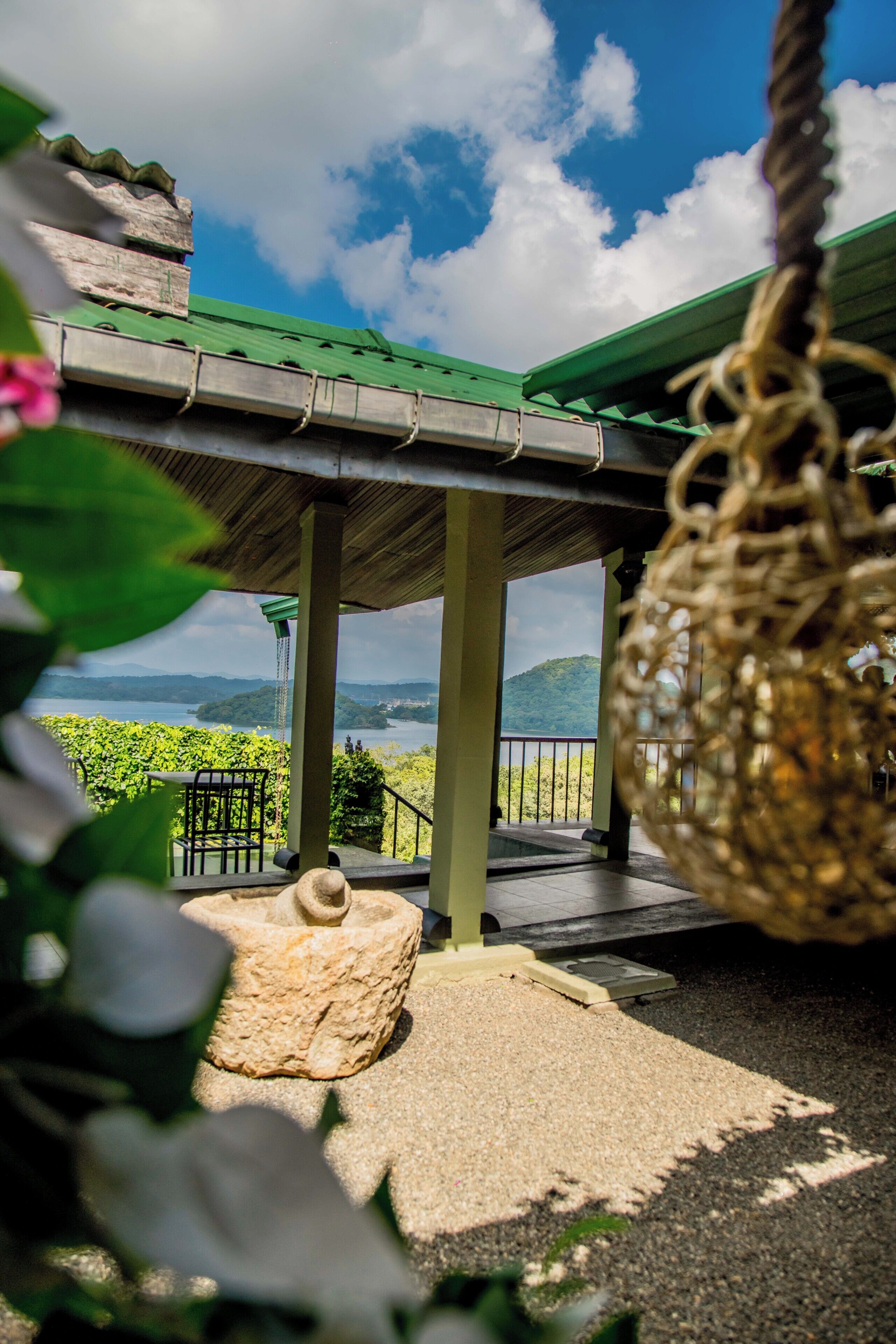 A covered outdoor area with a view of a lake and hills, featuring a swing, a stone bench, and a pergola.