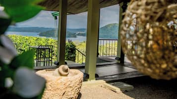 A covered outdoor area with a view of a lake and hills, featuring a swing, a stone bench, and a pergola.