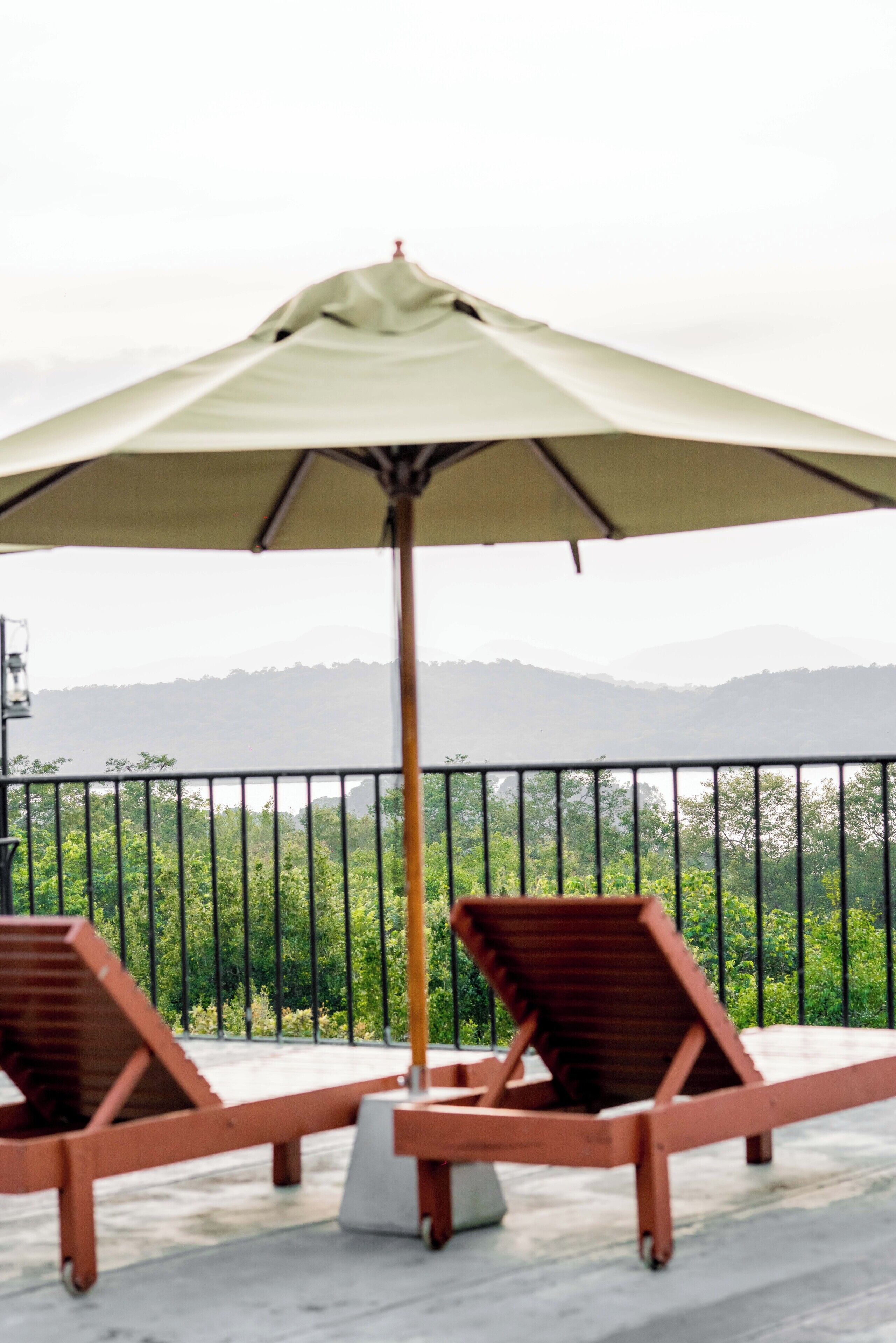 Two lounge chairs with a large umbrella, a railing, and a view of a mountainous landscape.