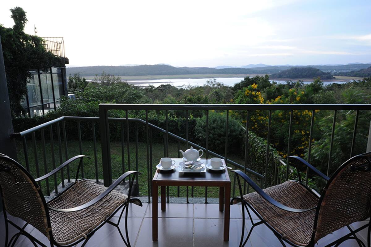 A balcony with wicker chairs and a table set for tea, overlooking a lake and hills.