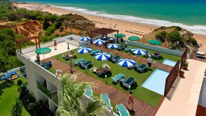 Un área de piscina en la azotea con sillones de descanso y sombrillas, con vistas a una playa y el océano.
