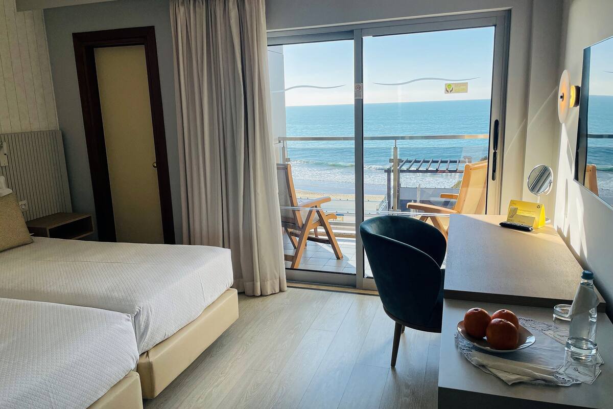 A hotel room with two beds, a desk with a chair, a table with oranges and a bottle, and a view of the sea through a sliding door.