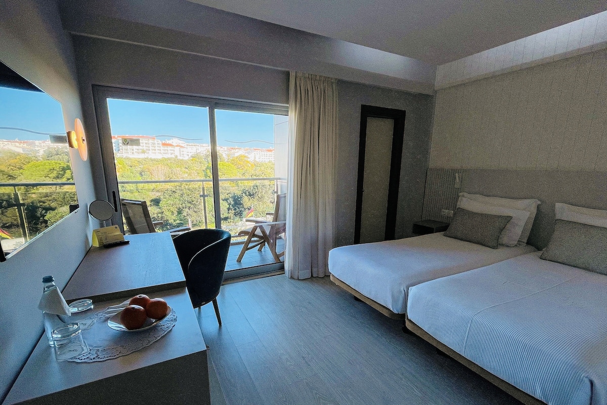 A hotel room with a large window, a bed, a desk with a chair, and a small table with a plate of oranges.