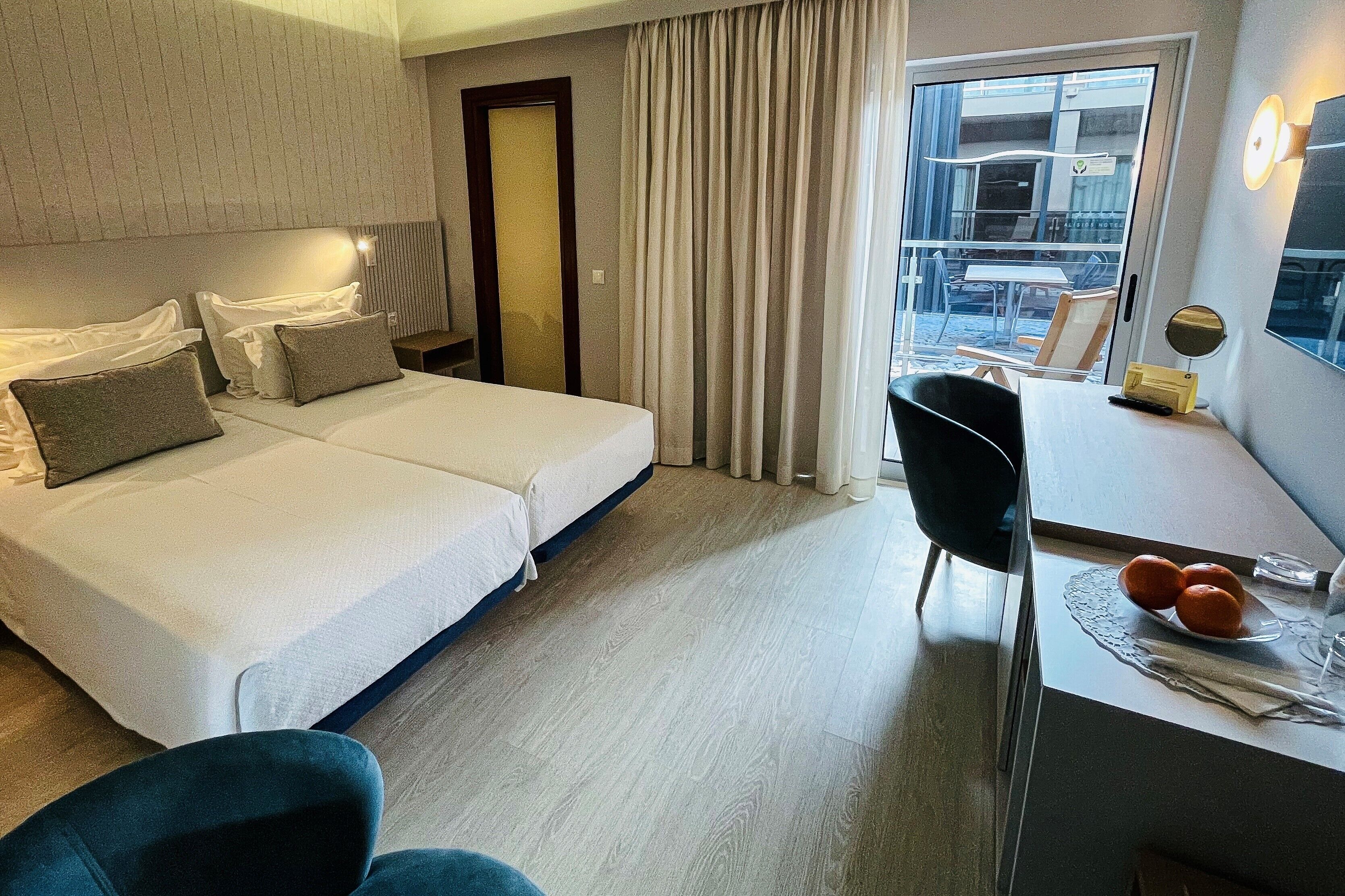 A hotel room with two beds, a desk with a chair, a small table with oranges, and a view of the outside through a window with curtains.