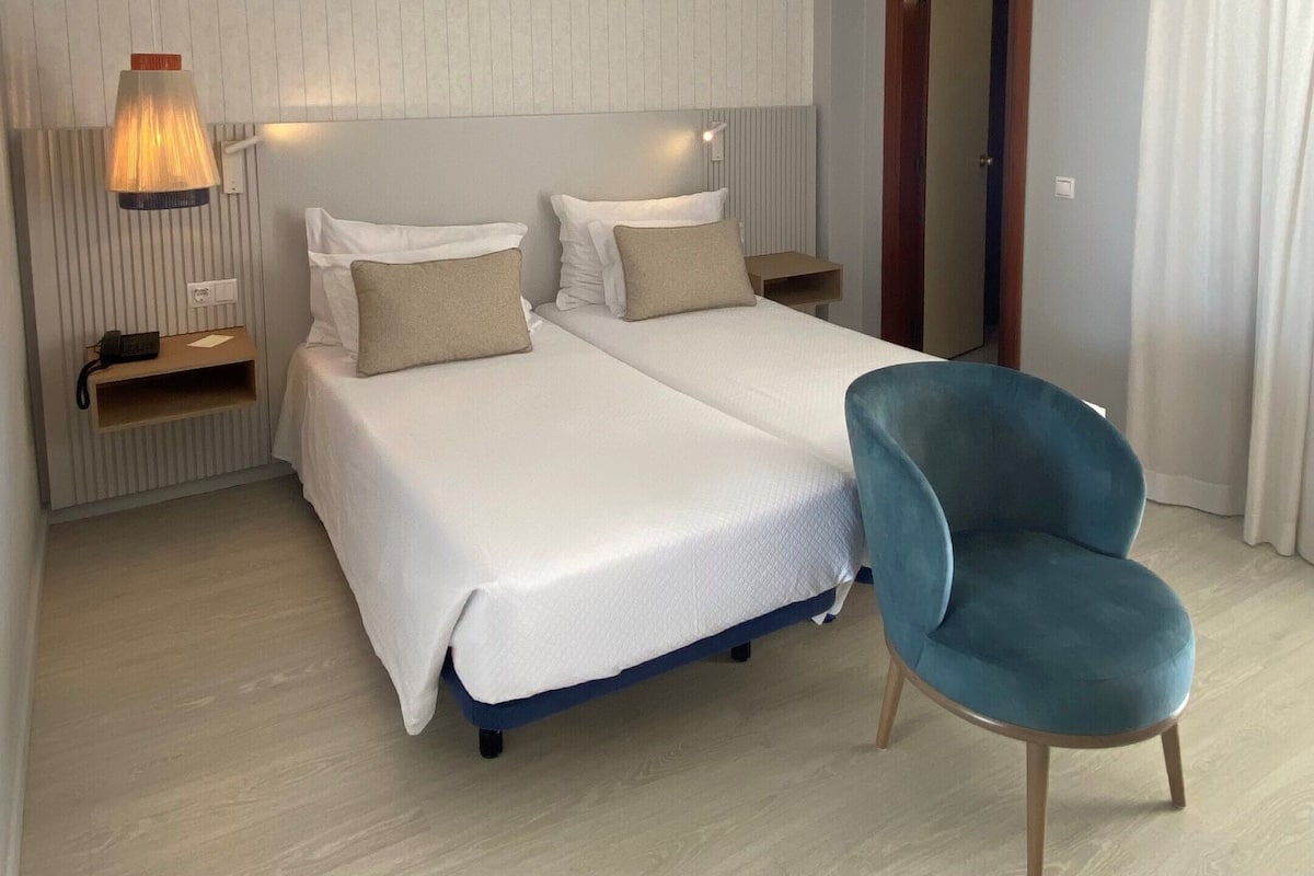 A hotel room with a double bed, a bedside table with a lamp, a chair, and a door leading to another room.