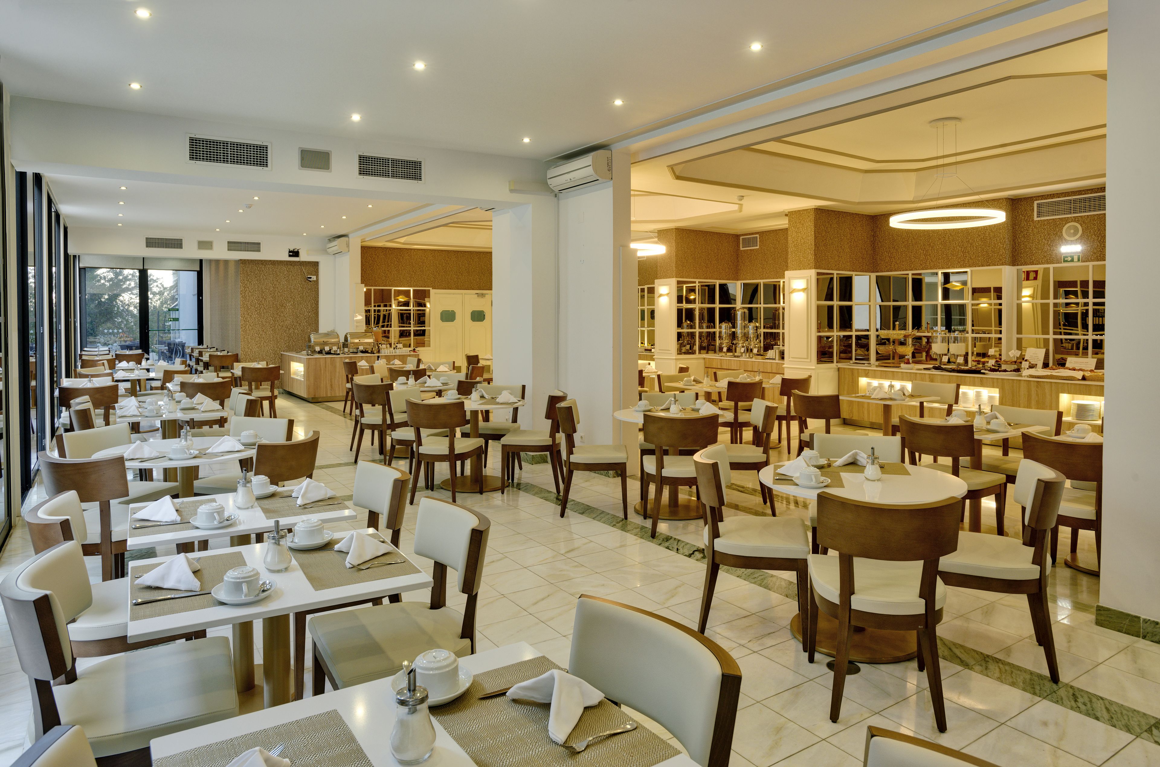 Un vaste espace de restauration d’hôtel, doté de nombreuses tables et chaises, de grandes fenêtres et d’un intérieur bien éclairé.