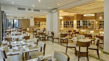 Un vaste espace de restauration d’hôtel, doté de nombreuses tables et chaises, de grandes fenêtres et d’un intérieur bien éclairé.
