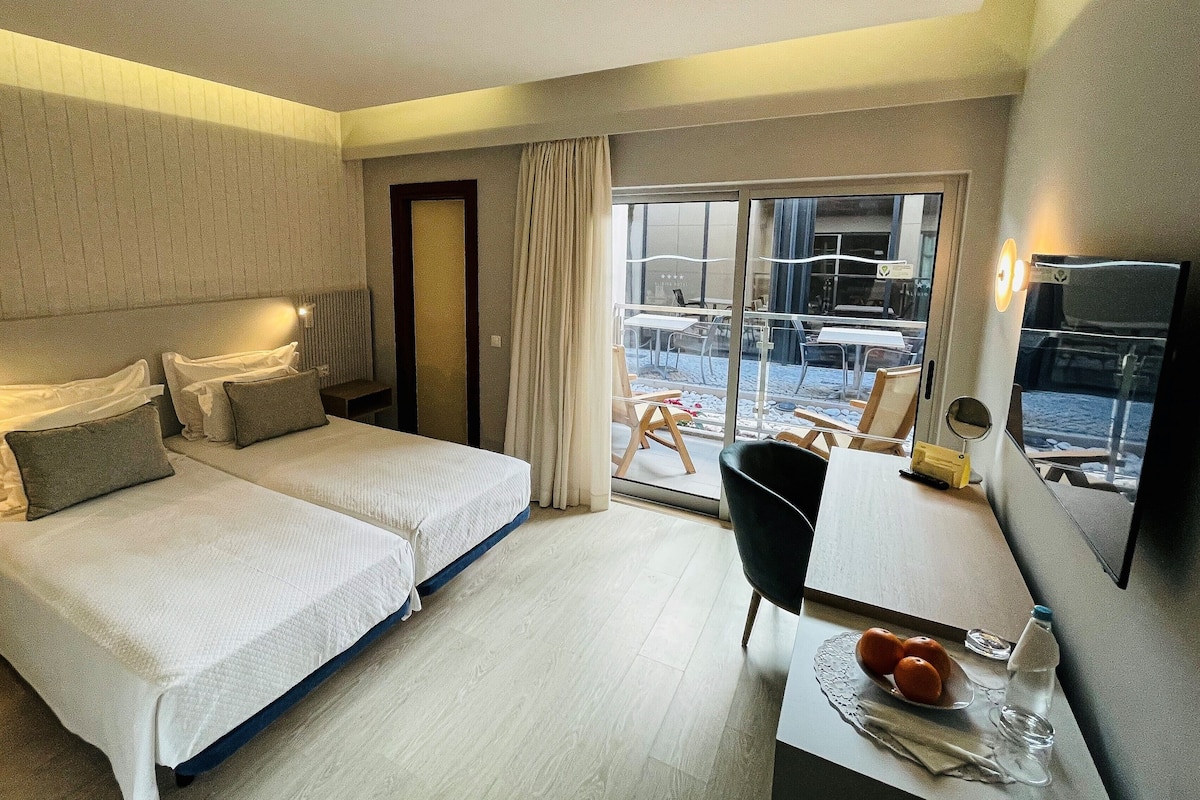 A hotel room with two beds, a desk with a chair, a TV, and a balcony with outdoor seating.