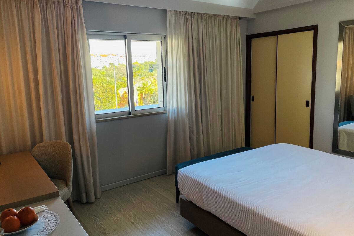 A hotel room with a bed, a desk, a chair, a window with curtains, and a sliding door.