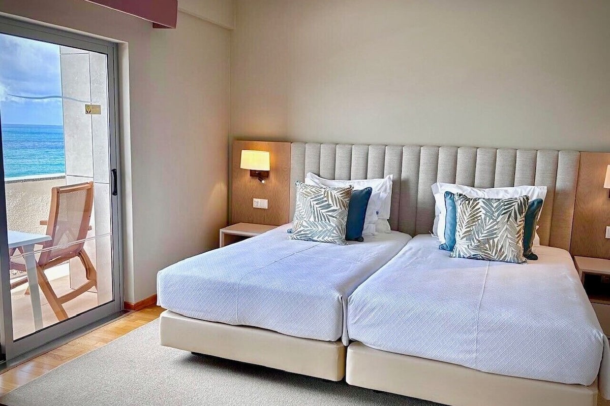 A double bed with two nightstands, a large window with a view of the ocean, and a chair by the window.
