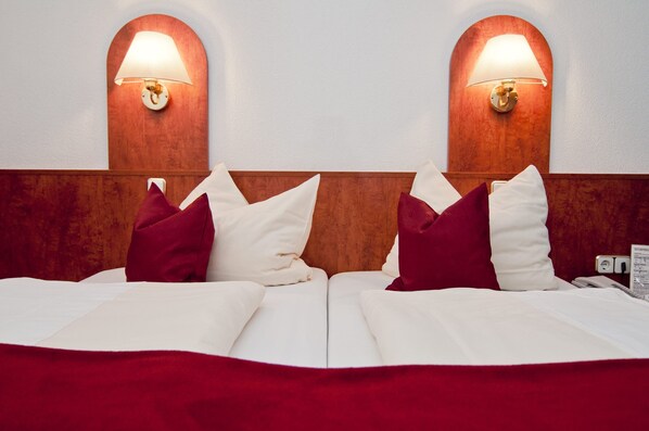 Uma cama de casal com roupa de cama branca e detalhes vermelhos, duas luminárias fixadas na parede e uma cabeceira de madeira.